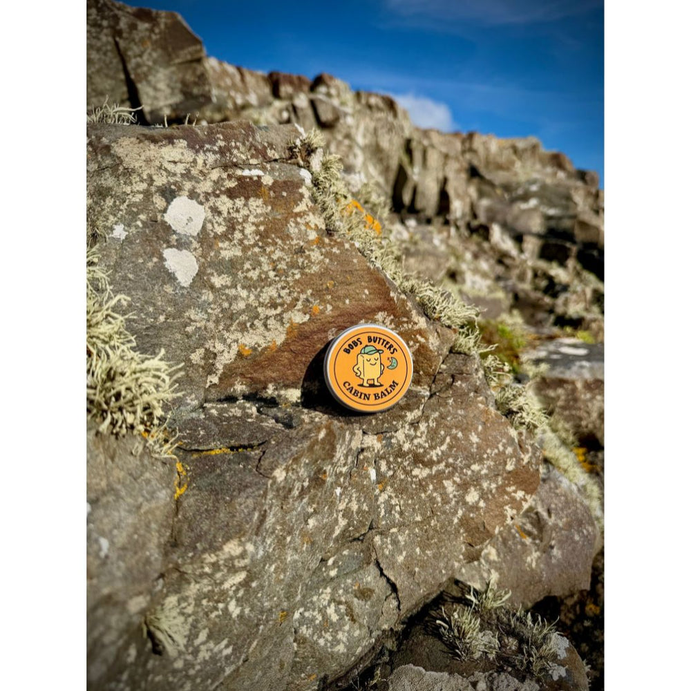 A round amber-colored butter balm tin sits on a mossy rock outdoors, with a rocky cliff and blue sky in the background.