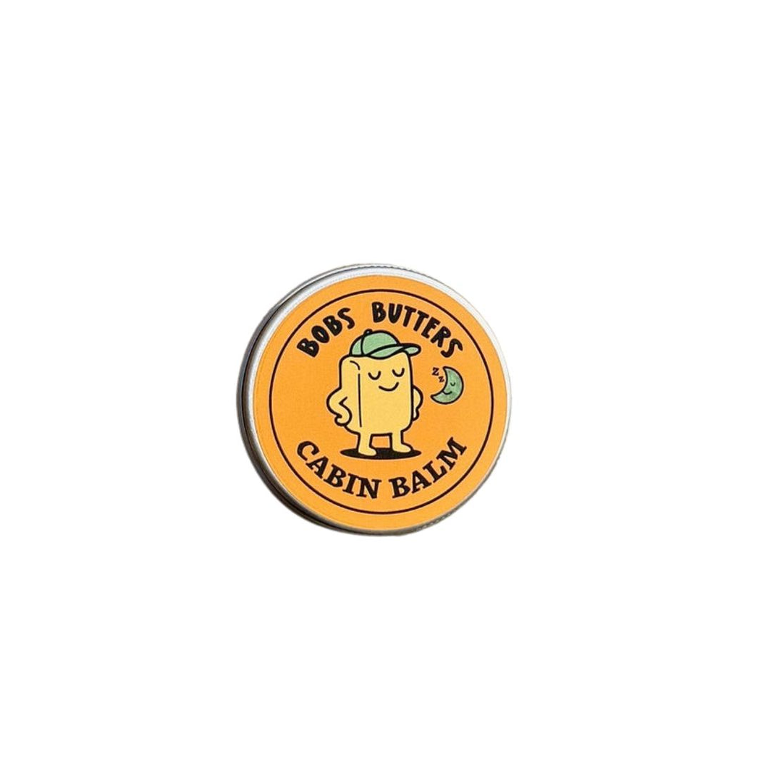 A round tin of Bob's Butters Cabin Balm with a bright orange label featuring a cartoon character and black text, set against a plain white background.