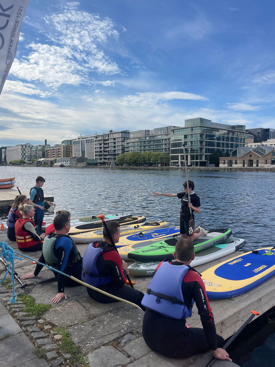 Group of people with paddleboards on a dock by a body of water with buildings in the background.